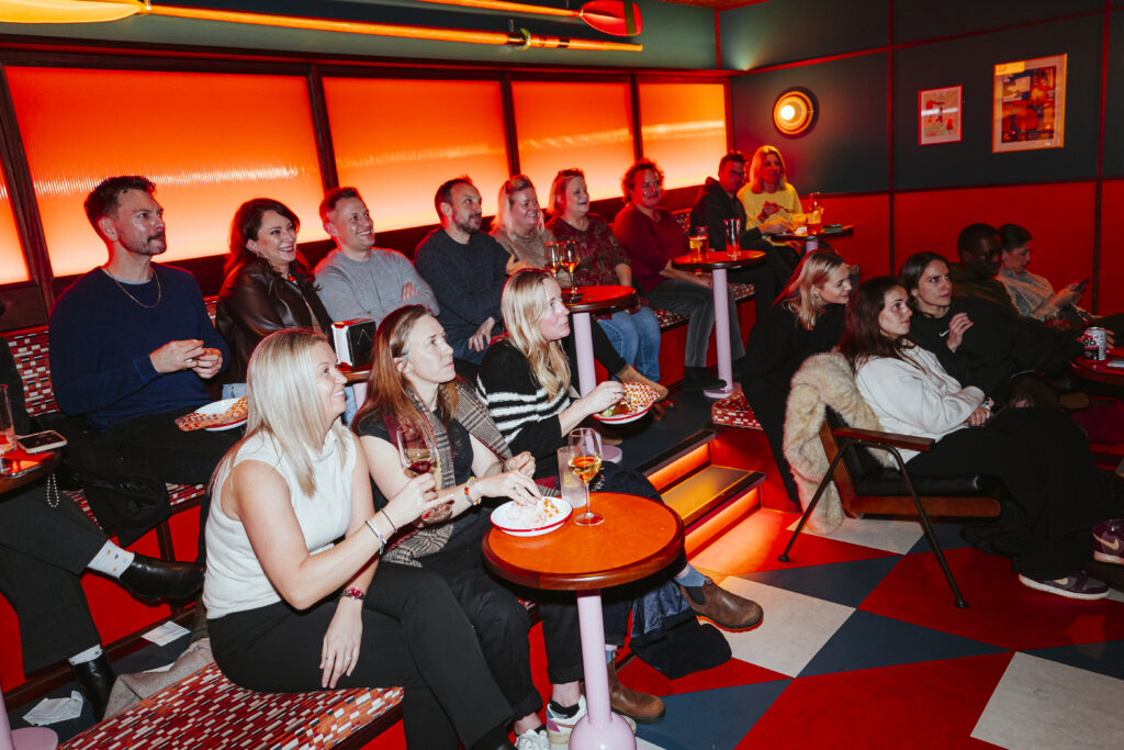 A group of people sit on tiered benches in a colourful, warmly lit room, watching something off-frame. With drinks and snacks in hand, they appear engaged and relaxed—a perfect scene if you want to book a private room for your next gathering.
