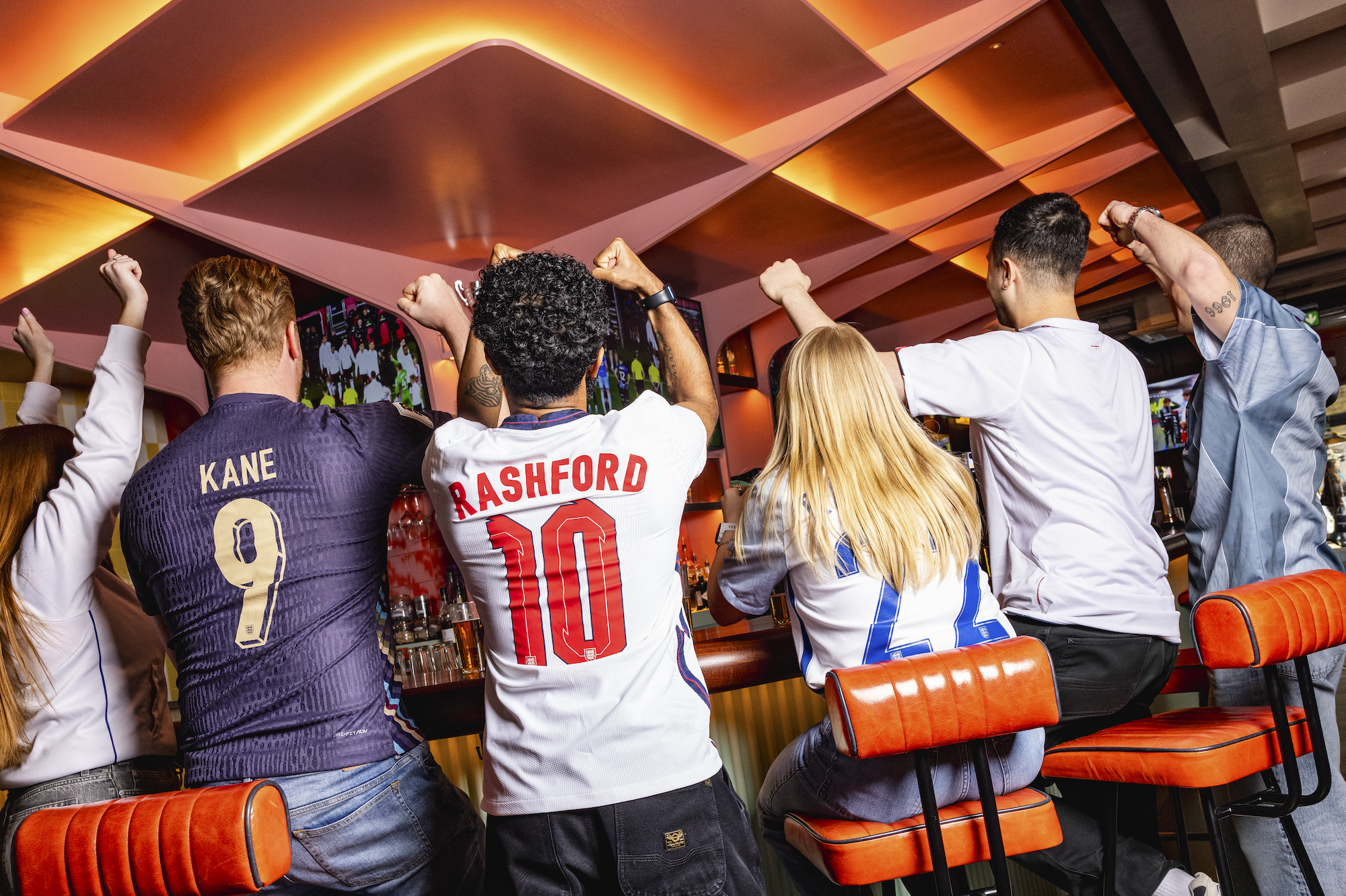 Five people sit on bar stools, cheering and watching a World Cup 2026 match on TV screens. They wear football shirts with names like Kane and Rashford. The scene is lively, filled with excitement and orange lighting overhead.