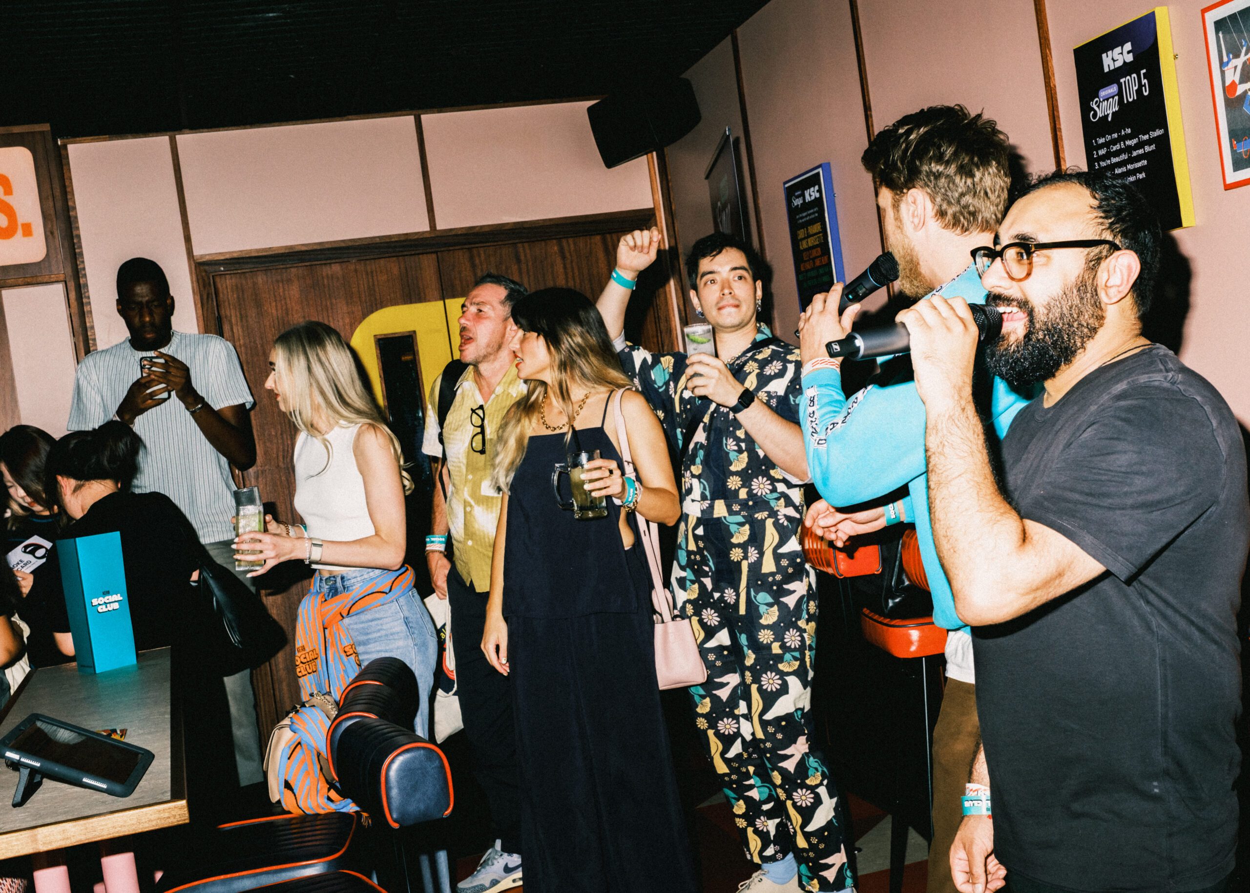A group of people in casual clothing enjoy drinks and socialise in a lively bar with colourful Christmas decor. One person holds a microphone, while others talk, laugh, and use their mobiles.
